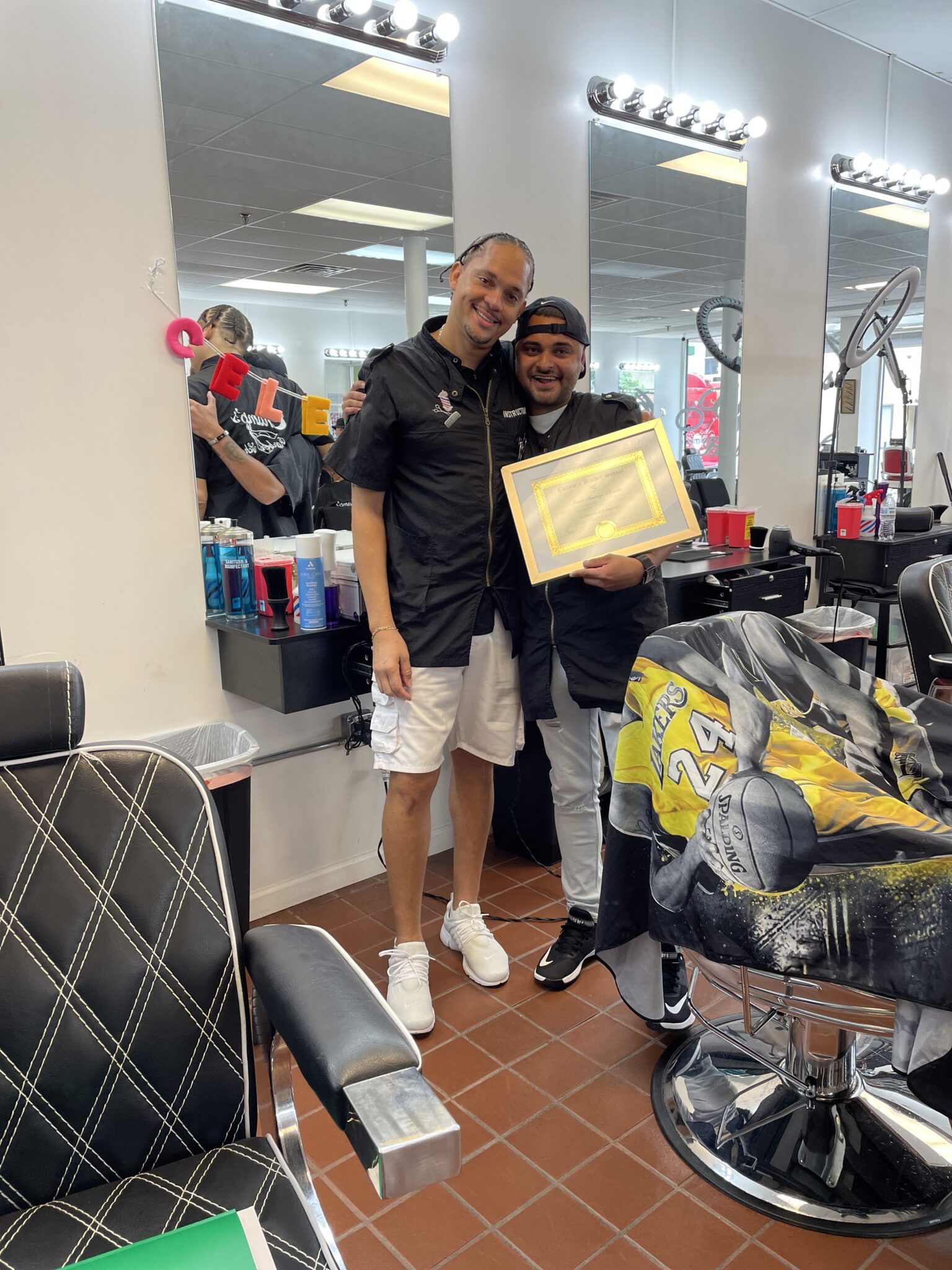 Two men smiling in a barbershop, one holding a certificate.